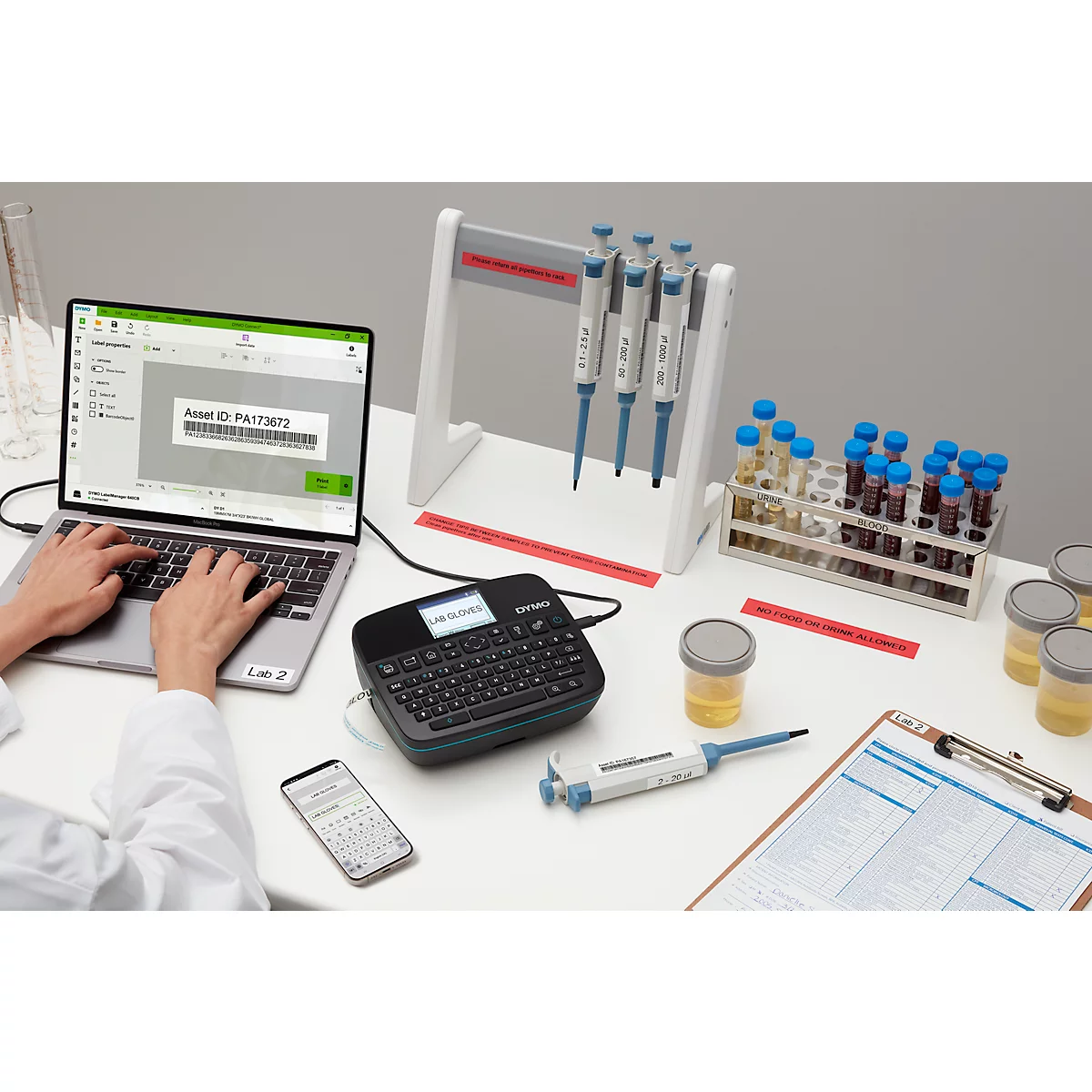 Eine Person tippt auf einem Laptop. Daneben befinden sich Etikettendrucker, Becher, Reagenzgläser und Pipetten.