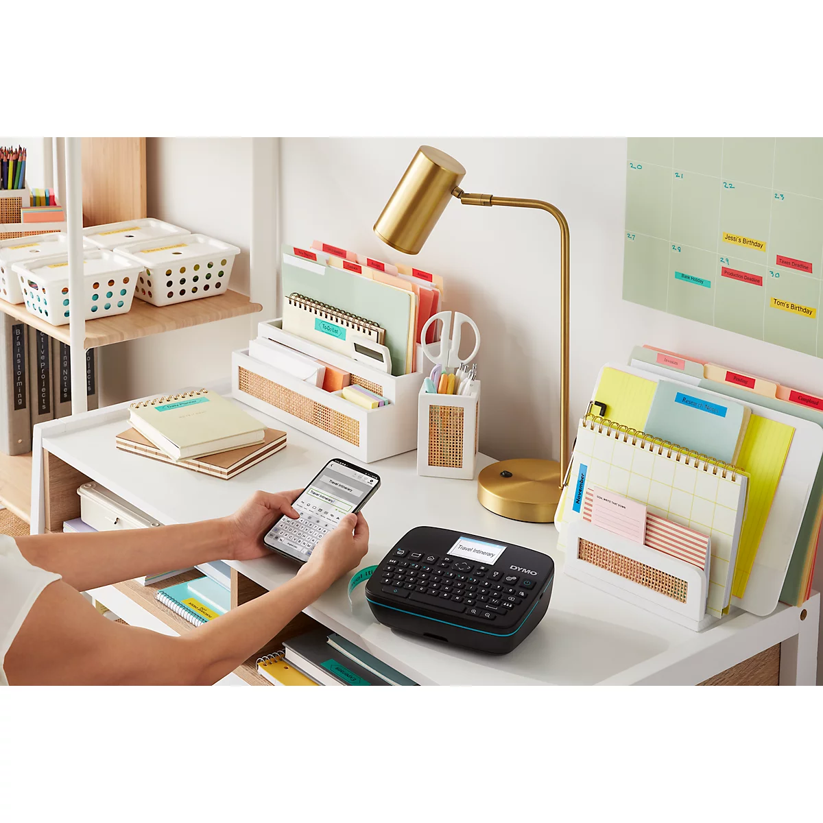 Een persoon die een labelmaker op een bureau gebruikt. Het bureau bevat notitieboekjes, pennen, ordners en een lamp.