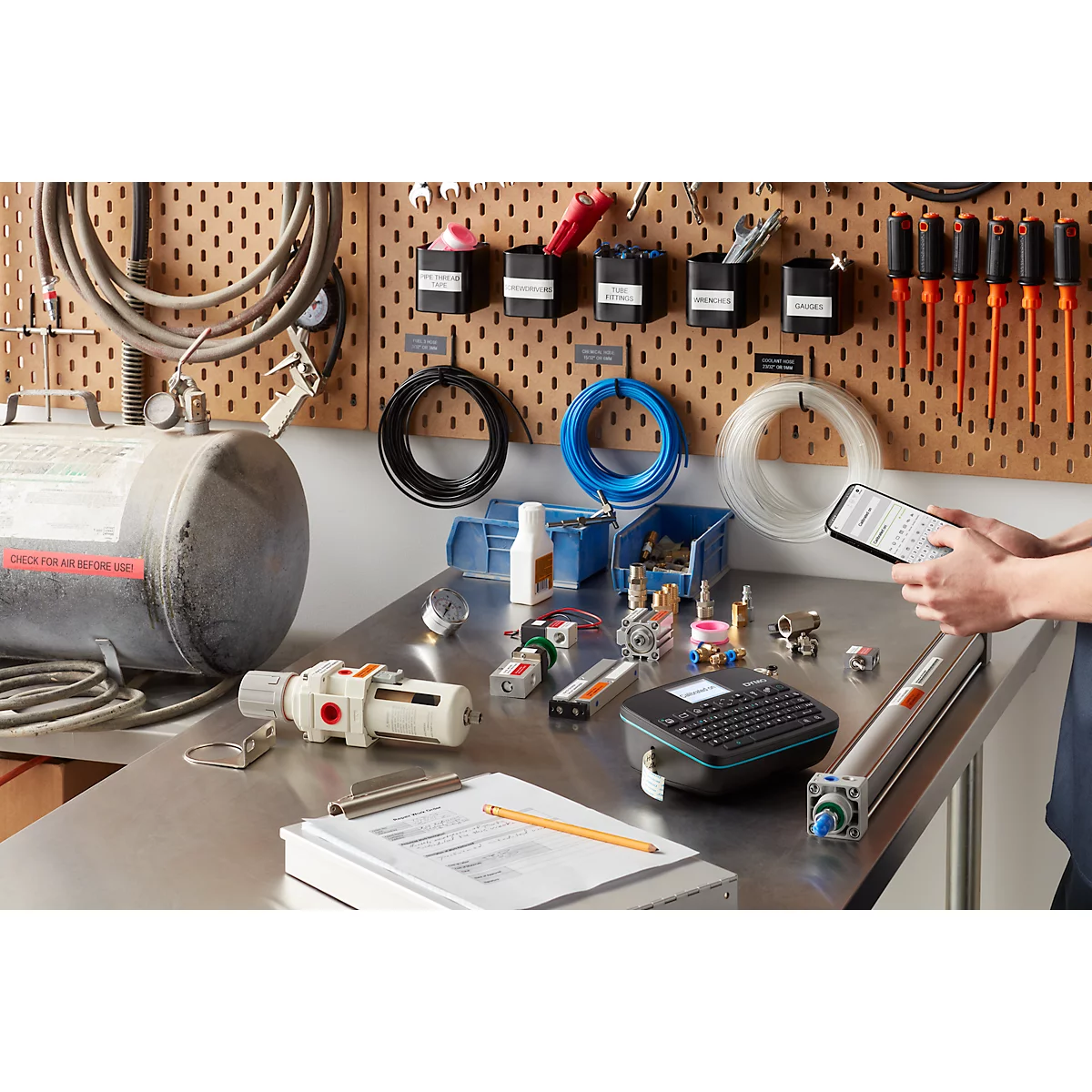 Werkbank met gereedschap, buizen, labelprinter en persoon die een tablet bedient.