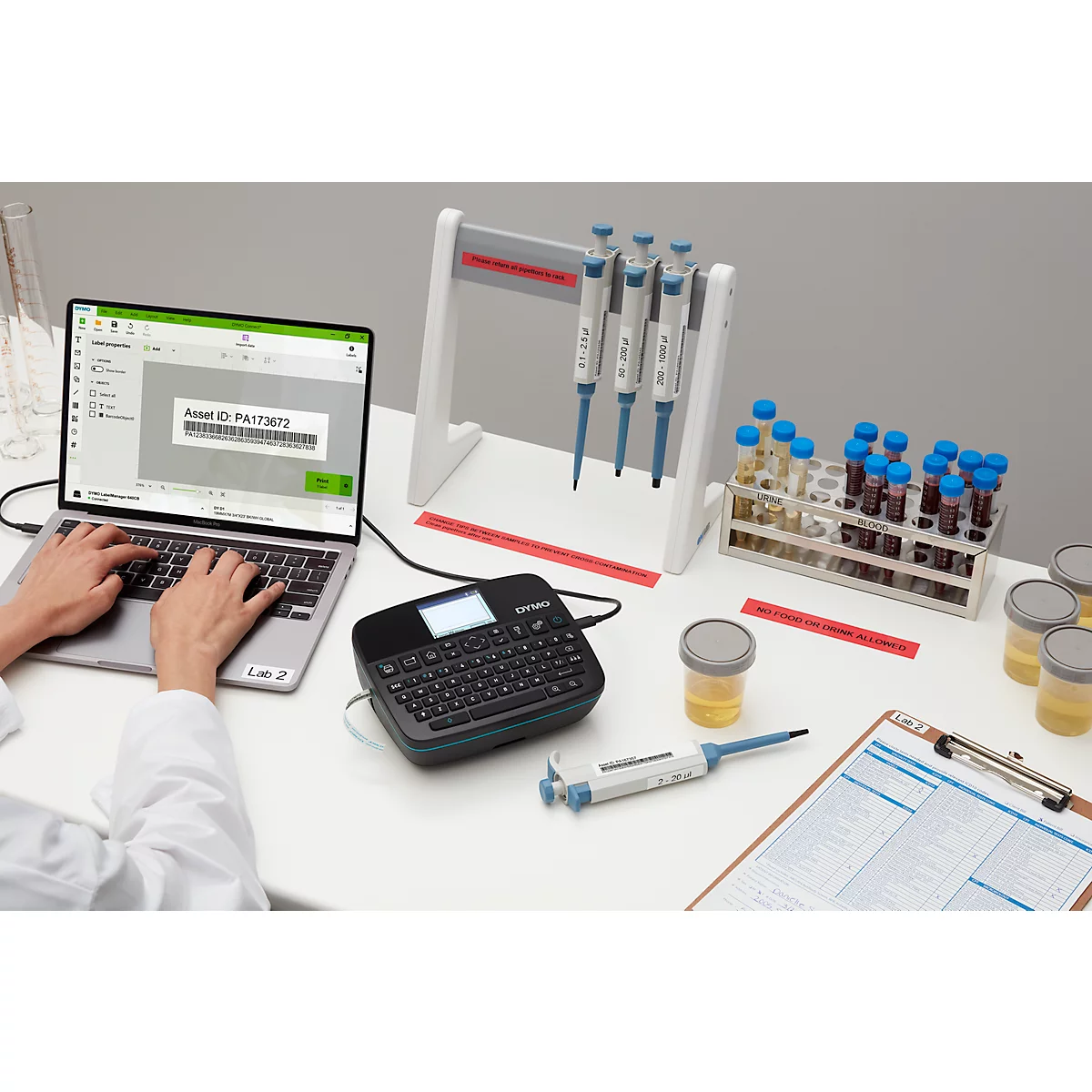 Personne tapant sur un ordinateur portable. A côté, du matériel de laboratoire, des pipettes, des tubes à essai, une imprimante d'étiquettes.