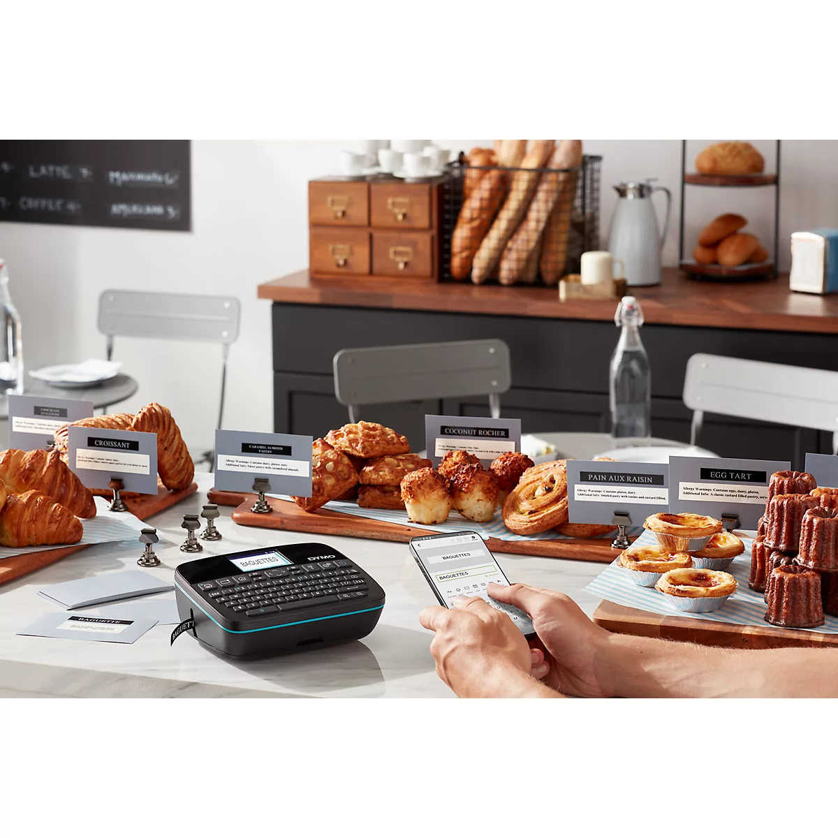 Personne utilisant un smartphone et une étiqueteuse. Des pâtisseries, des panneaux et du pain sont sur une table.