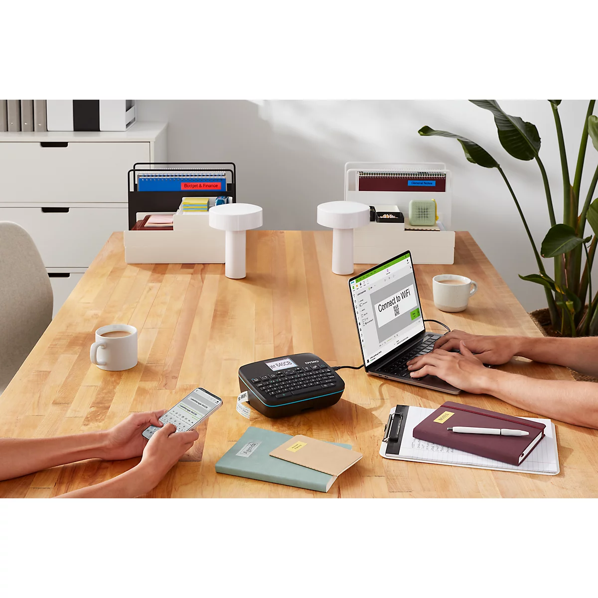 Scène de bureau: personne sur ordinateur portable, autres sur téléphone, étiqueteuse sur table, café, stylos.