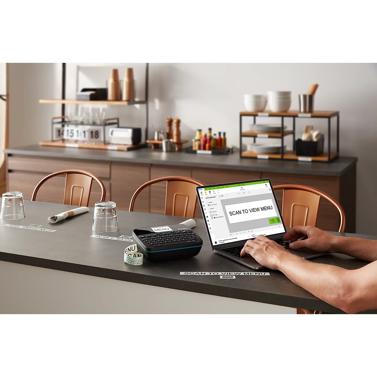 Personne tapant sur un ordinateur portable. L'écran affiche : "SCAN TO VIEW MENU". Sur la table : clavier, verres, ustensiles de cuisine.