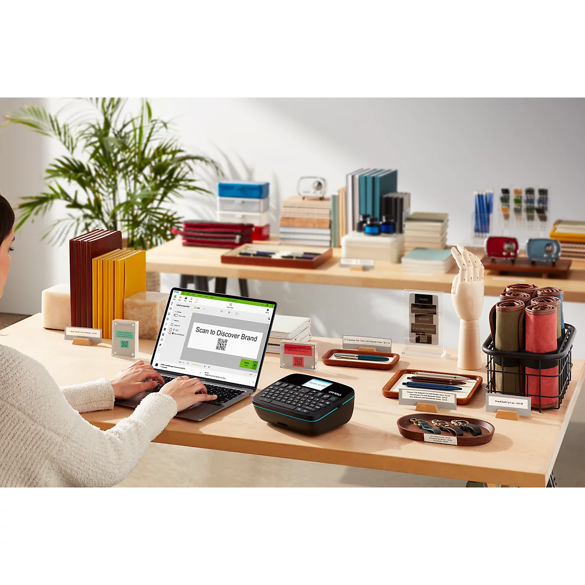 Une femme travaille sur un ordinateur portable avec une étiqueteuse sur une table. L'ordinateur portable affiche "Scan to Discover Brand" et un code QR.