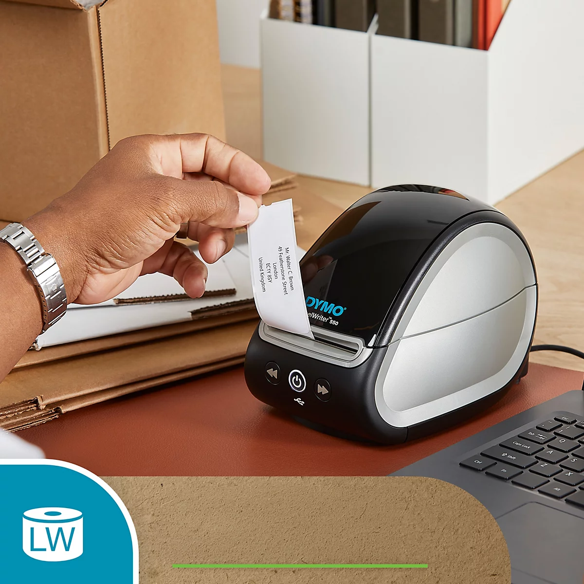 Eine Hand zieht ein Etikett aus einem schwarzen DYMO LabelWriter. Im Hintergrund sind Kartons und ein Laptop zu sehen.