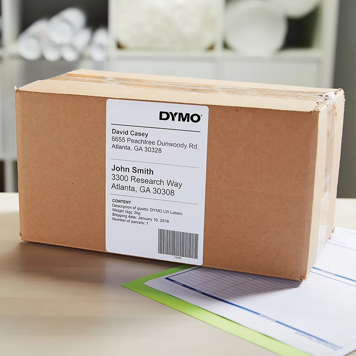 Brauner Karton mit DYMO-Etikett, Adressen und Barcode. Auf dem Tisch liegen weitere Dokumente.