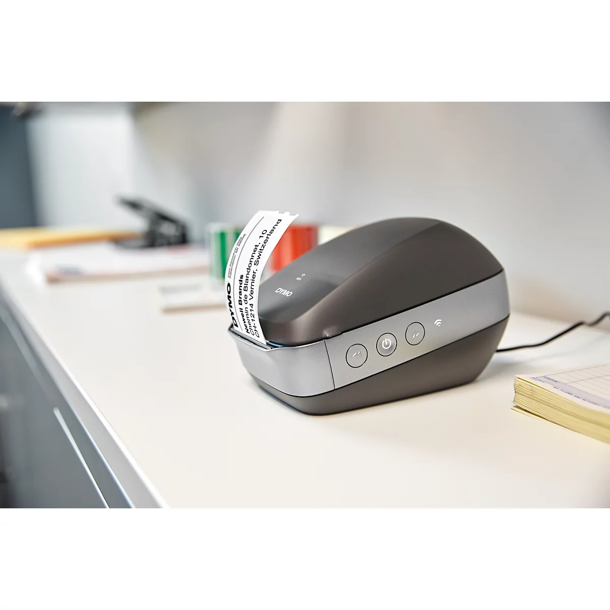 A label printer dispensing an address label sits on a white tabletop. Office supplies in the background.