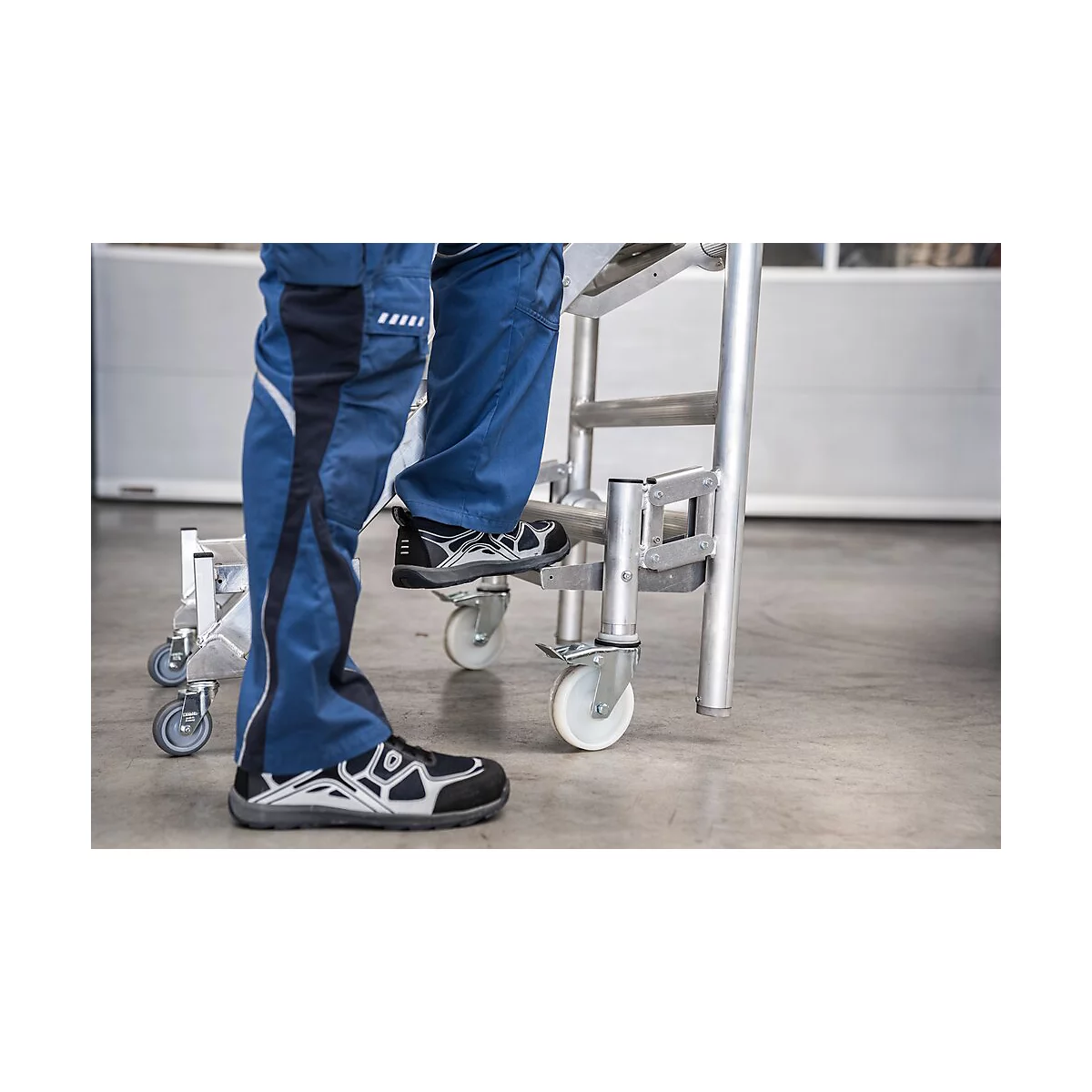 Personne debout sur une échelle à roulettes. Pantalon bleu, chaussures noires. Échelle en métal, fond gris.