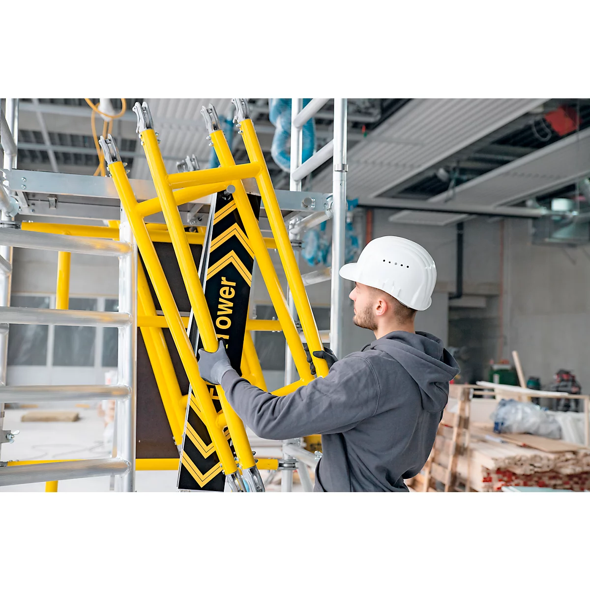 Un homme en vêtements de travail assemble un échafaudage jaune. L'échafaudage porte l'inscription "stower".