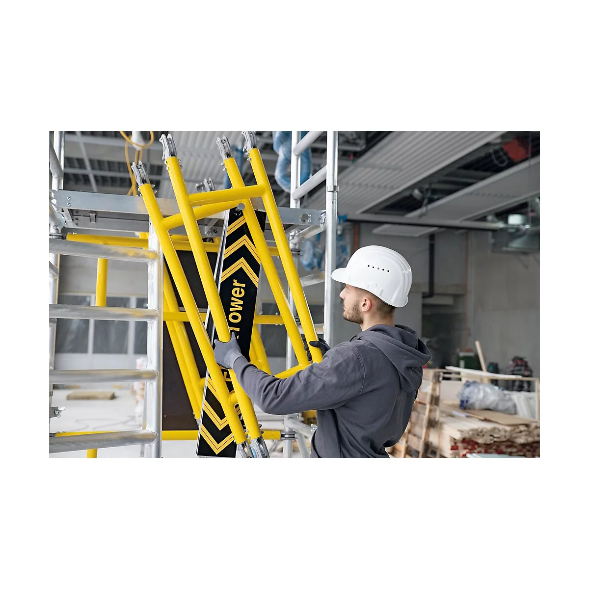 Un ouvrier avec un casque tient une partie d'échafaudage jaune. D'autres parties d'échafaudage et un chantier en arrière-plan.