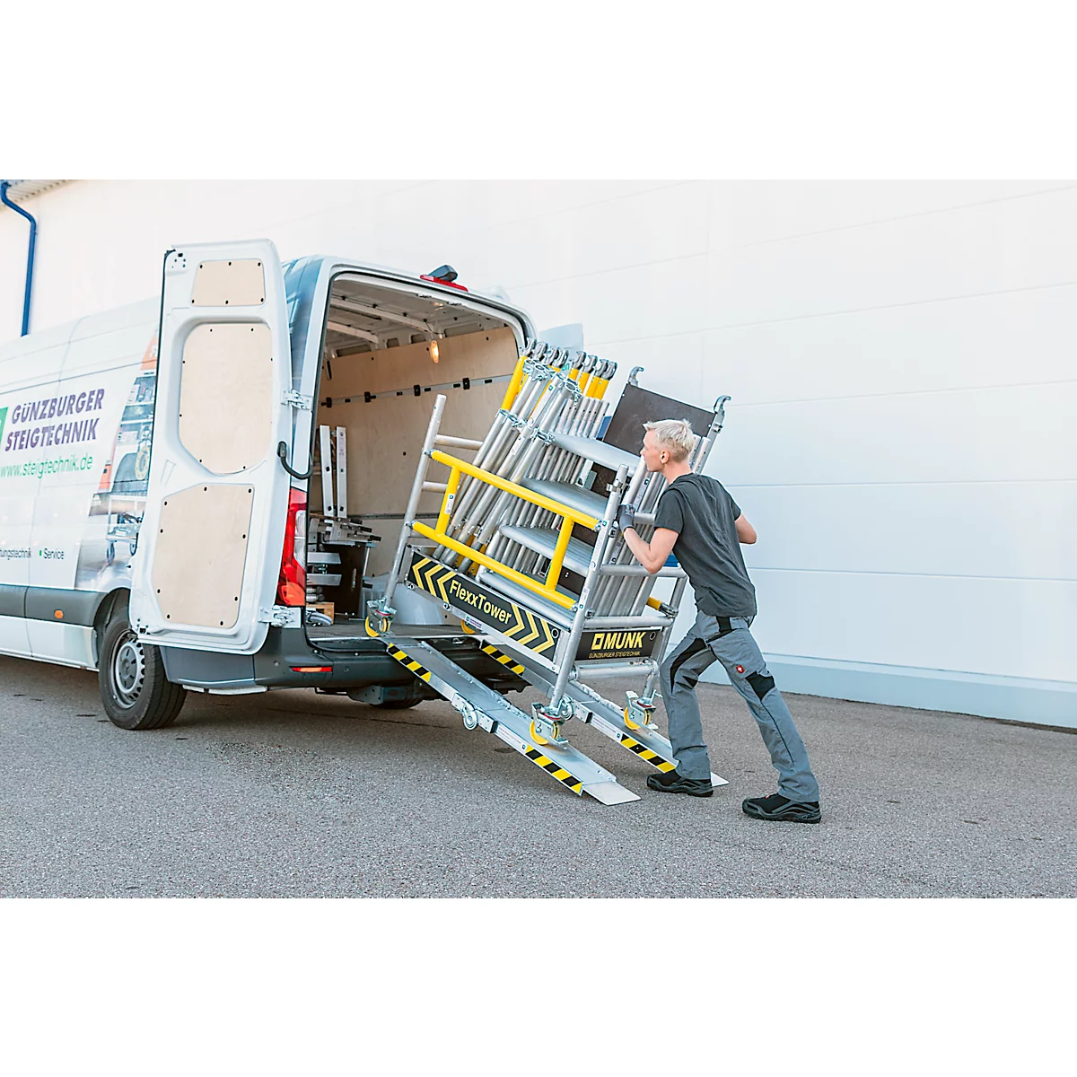 Un homme pousse un échafaudage hors d'une camionnette. Bandes d'avertissement jaunes et noires sur la rampe et l'échafaudage. Logo: DÜNZBURGER STEIGTECHNIK.