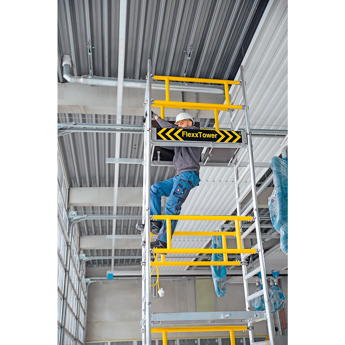 Un ouvrier sur un échafaudage assemble quelque chose au plafond. FlexxTower est sur l'échafaudage. Garde-corps jaunes.