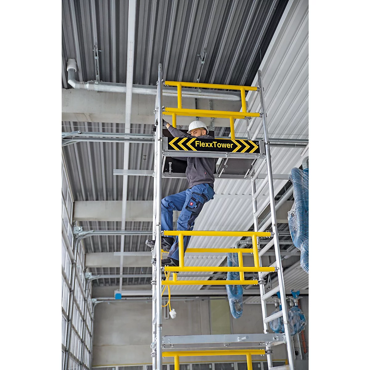 Un homme sur un échafaudage monte quelque chose au plafond. L'échafaudage a des garde-corps de sécurité jaunes. Sur l'échafaudage, il est écrit "FlexxTower".