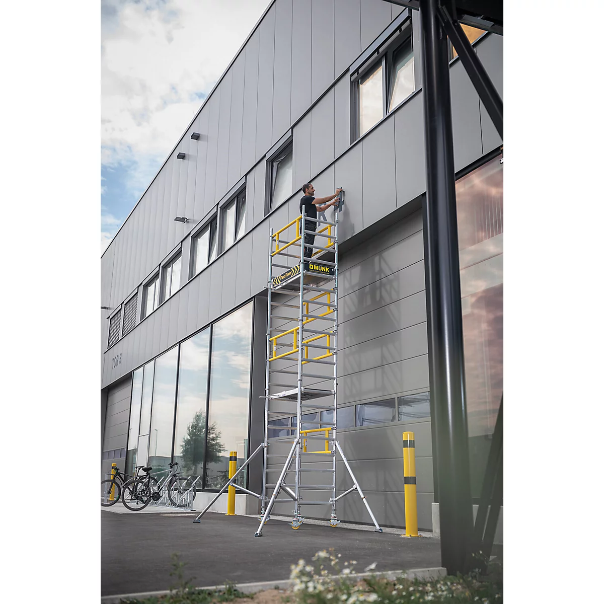Homme sur une échelle d'échafaudage, travaillant sur une façade de bâtiment. Structure d'échafaudage jaune et argentée, bâtiment gris avec des fenêtres en verre.