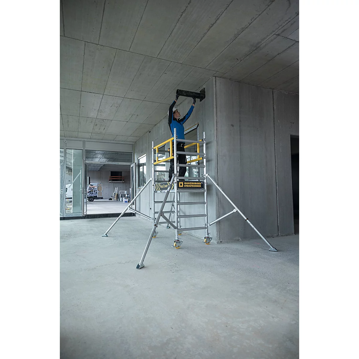 Une personne se tient sur un échafaudage et fixe un objet à un mur en béton. L'échafaudage a des éléments jaunes et se trouve dans un bâtiment en construction.