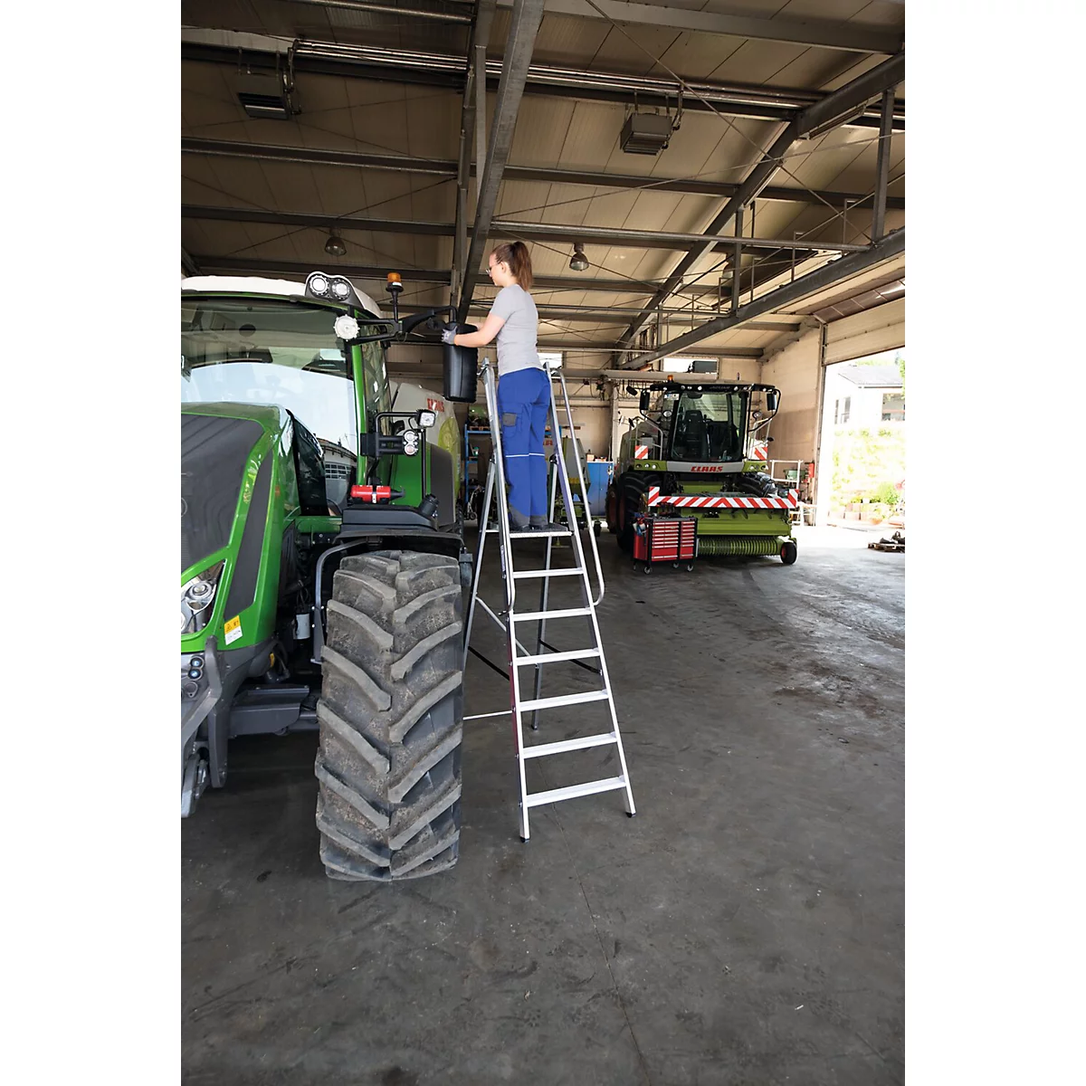 Une femme debout sur une échelle travaille sur un tracteur dans un atelier. En arrière-plan, il y a un autre tracteur.
