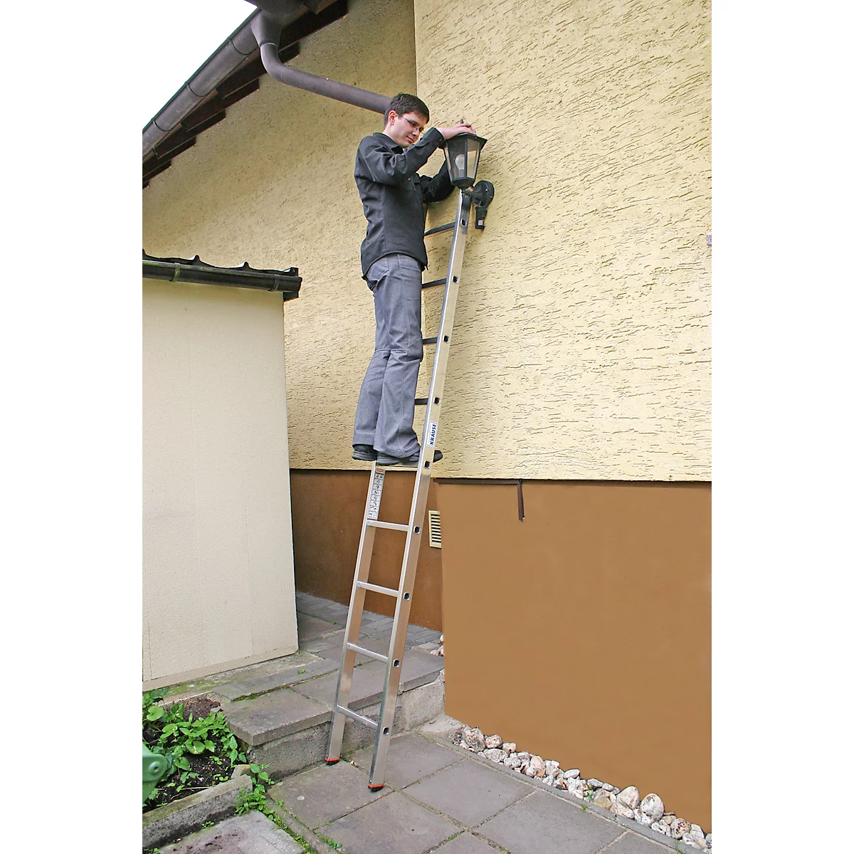Un homme sur une échelle fixe une lampe au mur de la maison. Mur de la maison beige, marron. Gouttière, trottoir, arbustes et gravier.