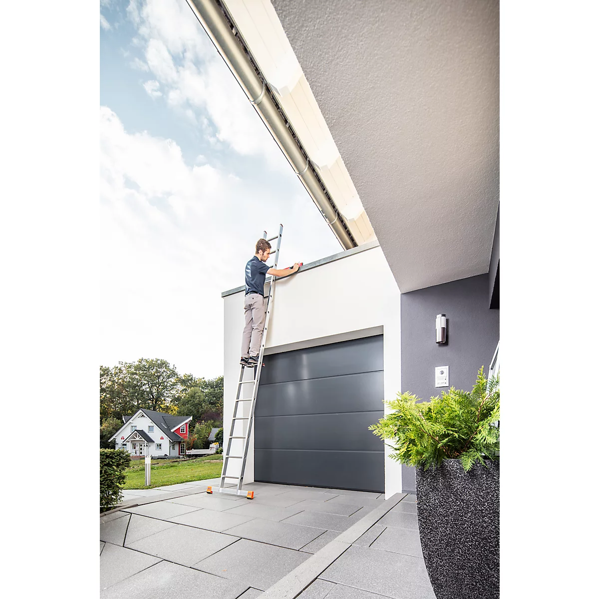 Un homme est sur une échelle près d'un garage. Il travaille sur la gouttière, devant un ciel bleu.