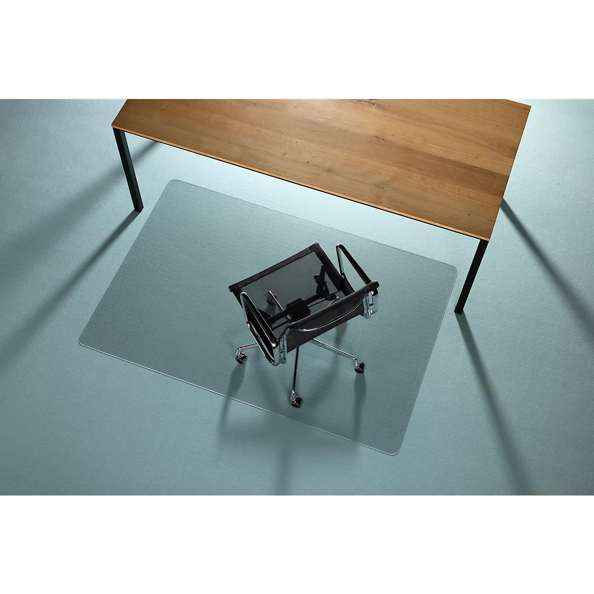 Silla de oficina sobre alfombrilla protectora de suelo transparente bajo mesa de madera. Superficie azul.