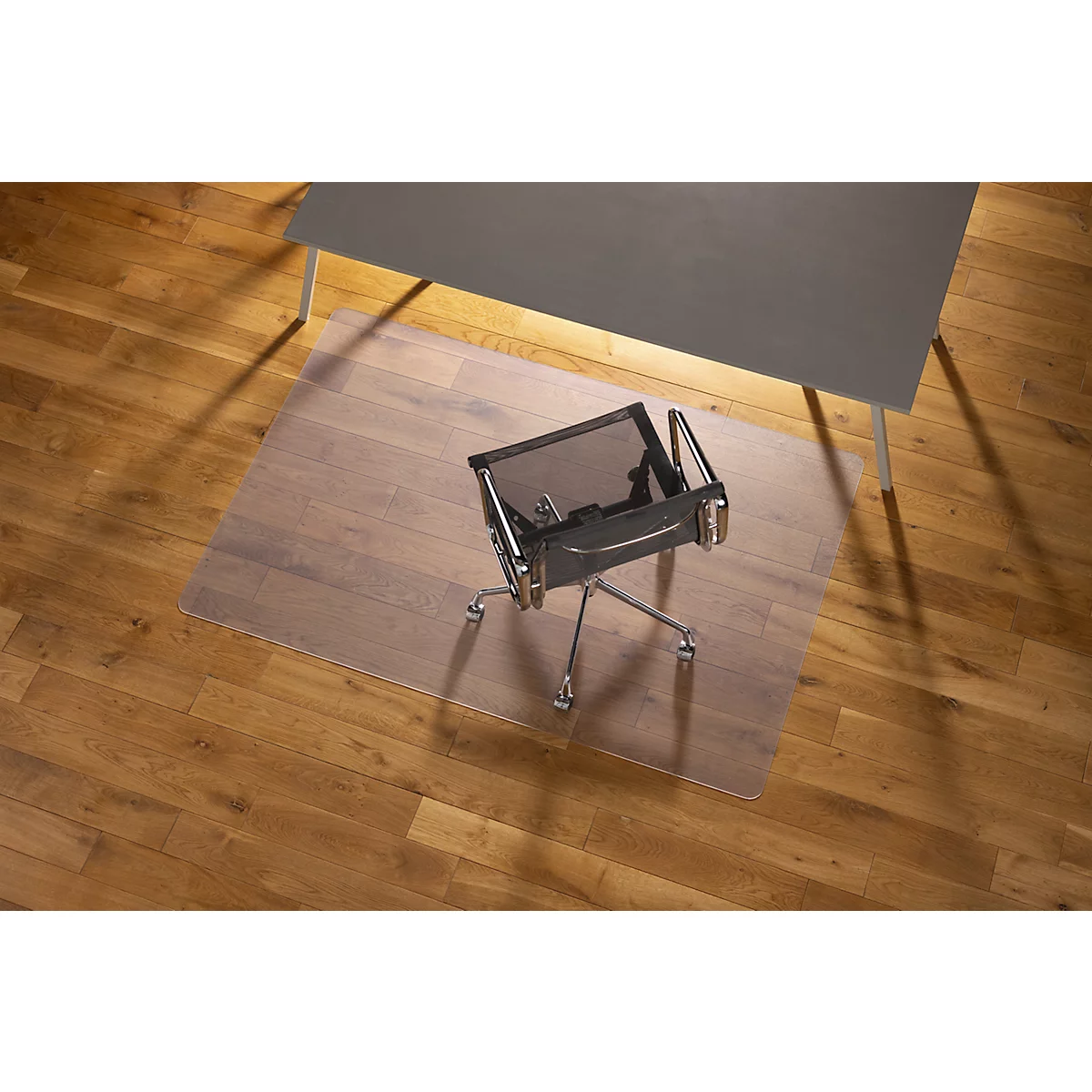 Silla de oficina sobre una alfombrilla transparente para escritorio en un piso de madera debajo de una mesa gris.