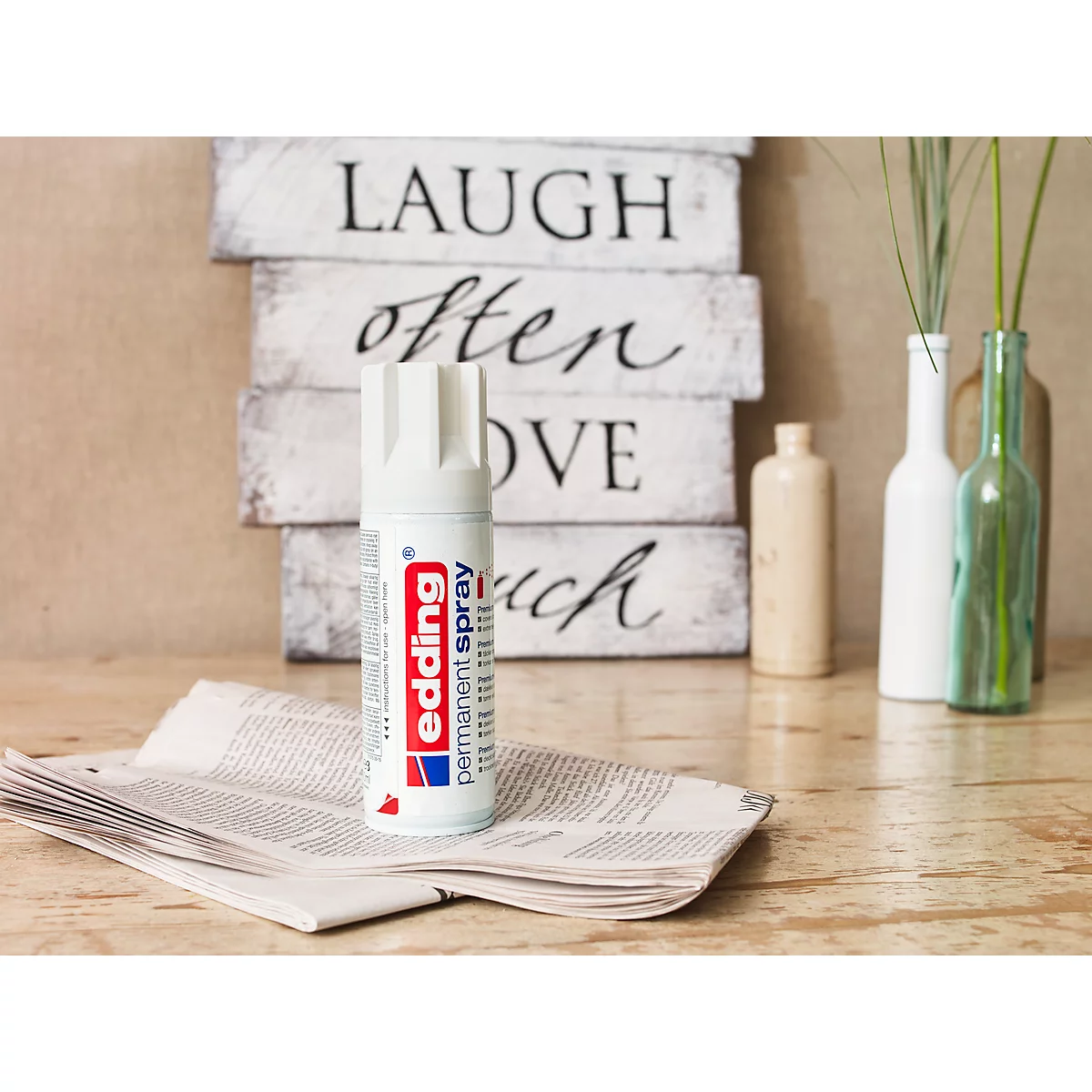 Een witte Edding permanente spuitbus staat op een krant, op de achtergrond houten borden met spreuken.