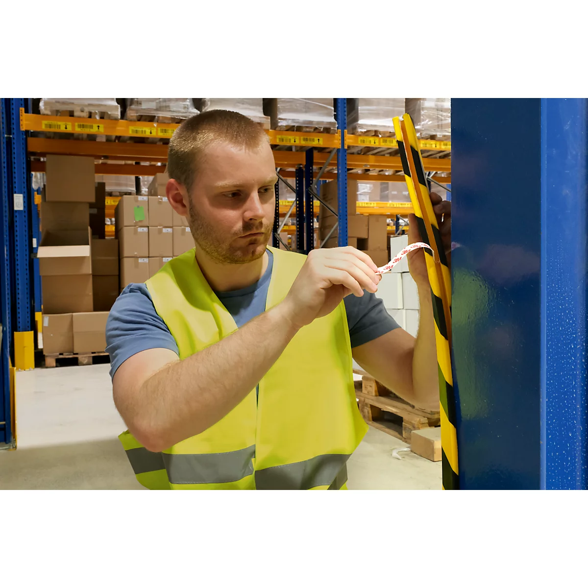 A man in a yellow safety vest attaches a tape to a warehouse pole. In the background, shelves with boxes are visible.