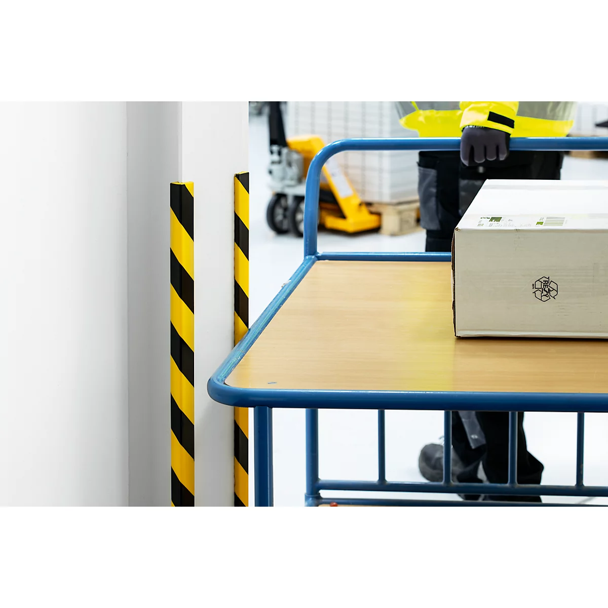 A worker pushes a cart with a cardboard box. Yellow and black warning poles, gray floor.