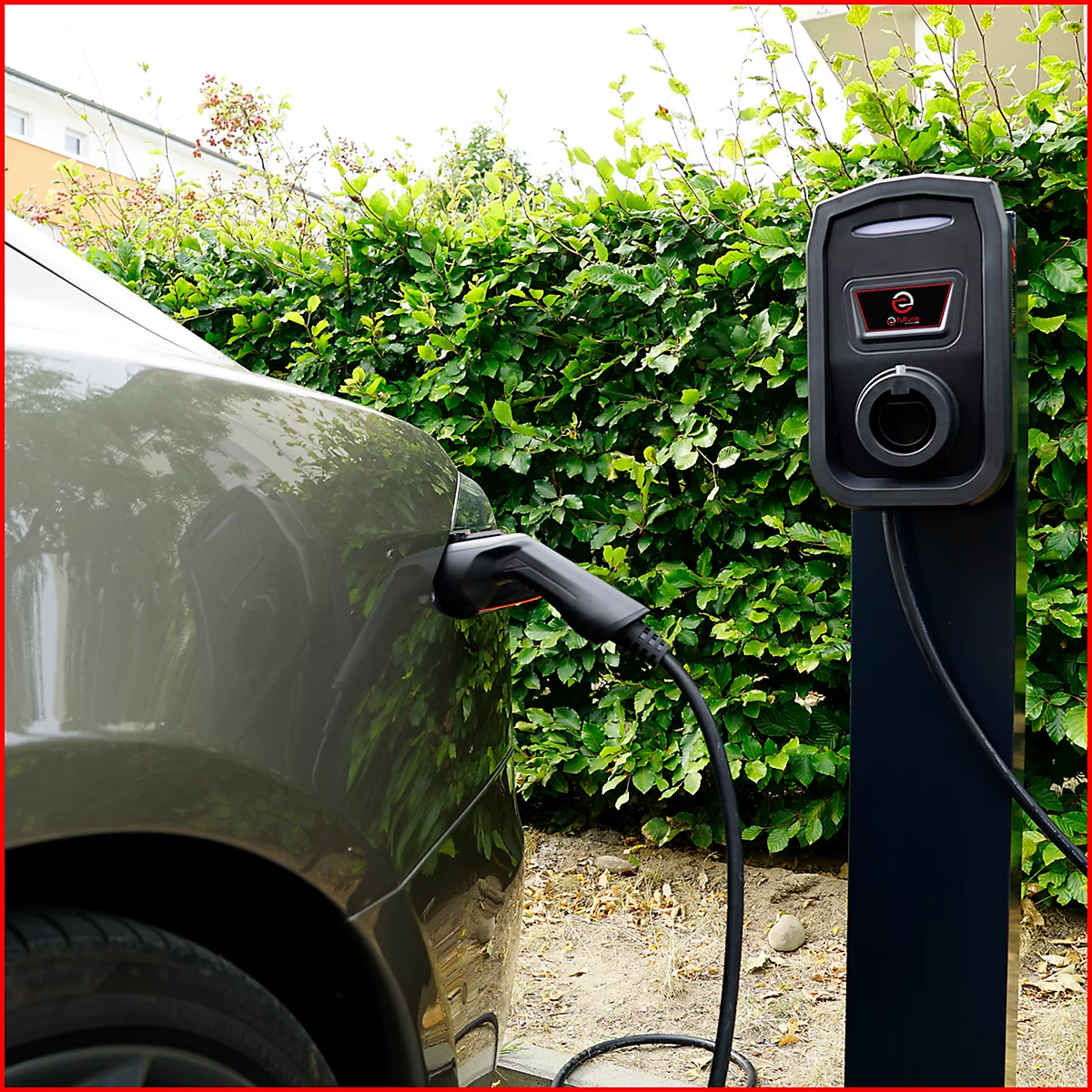 Graues Auto lädt an einer Ladestation. Schwarzes Kabel verbindet Auto und Ladestation. Im Hintergrund Hecke.