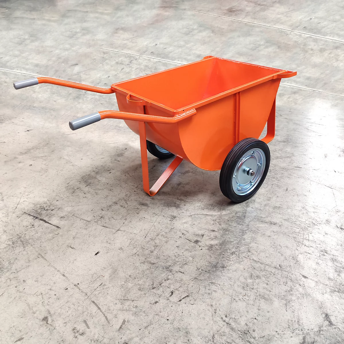 Eine orangefarbene Schubkarre mit zwei Rädern steht auf einem Betonboden. Die Griffe sind grau.