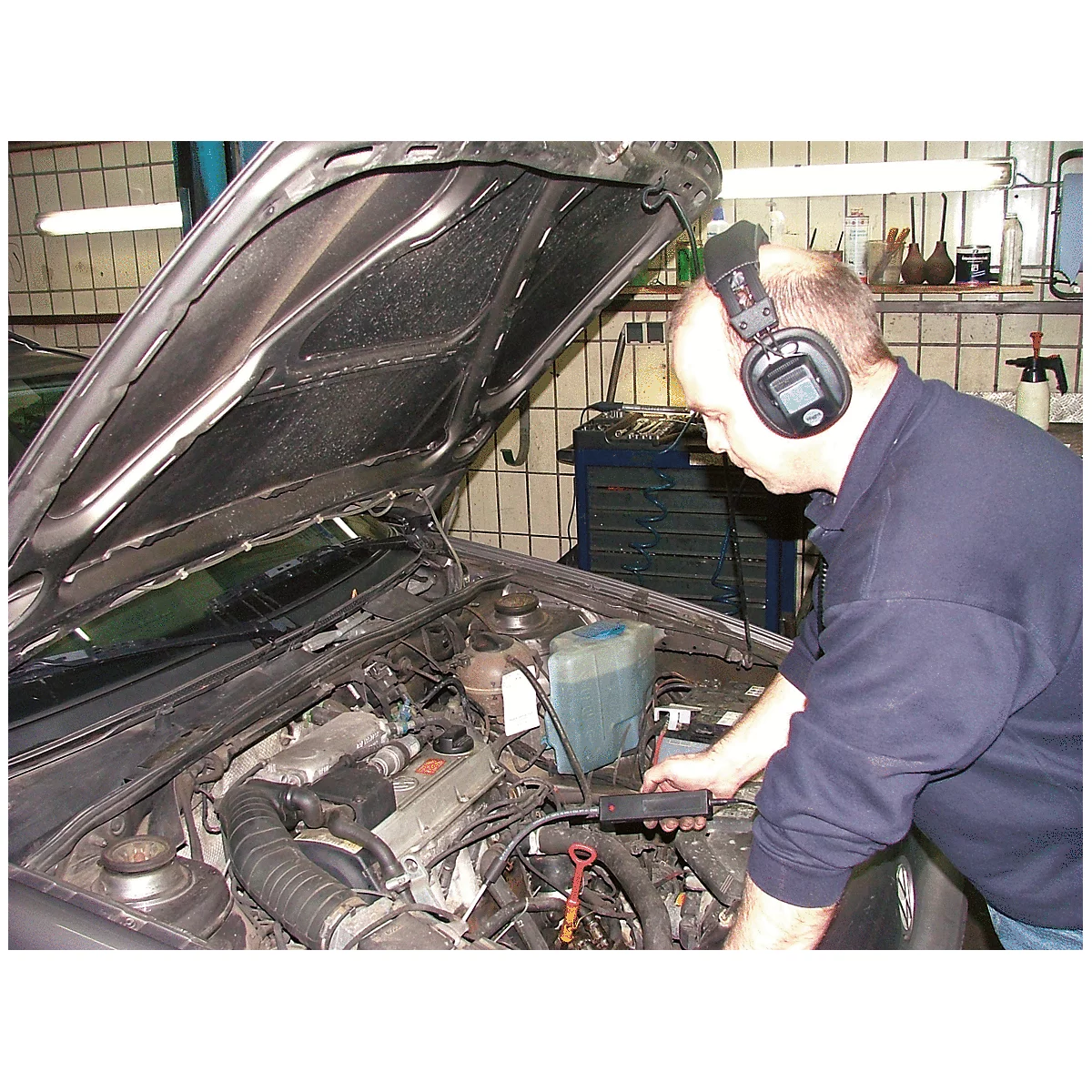 Ein Mechaniker mit Kopfhörern prüft einen Motorraum. Die Motorhaube ist geöffnet. Im Hintergrund Werkzeuge.