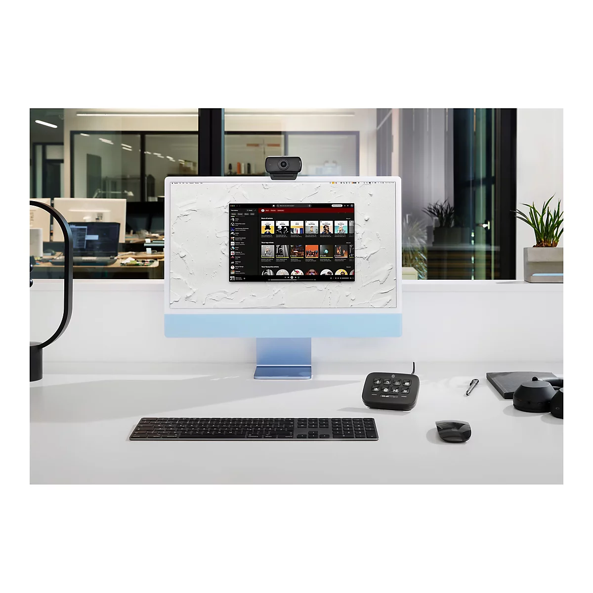 Ein modernes Büro-Setup mit einem blauen iMac, Webcam, Tastatur, Maus und anderen Arbeitsgeräten auf einem weißen Schreibtisch. Der Bildschirm zeigt eine Musik-Streaming-Oberfläche.