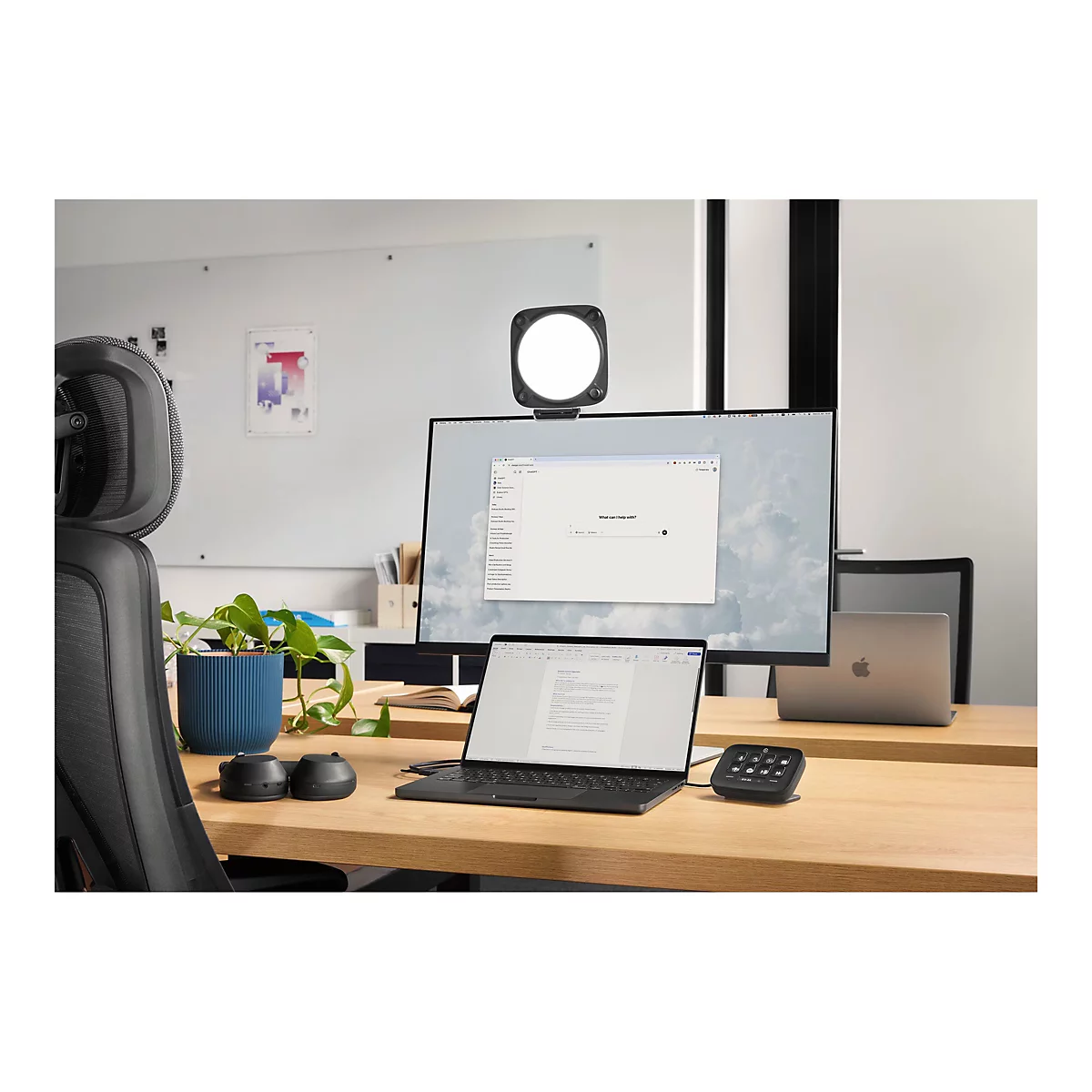 Schreibtisch mit Monitor, Laptop, Licht und Bedienfeld. Im Hintergrund Bürostuhl und Topf mit Pflanze.
