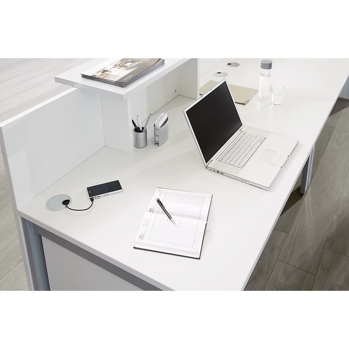 Aufgeräumter, weißer Schreibtisch mit Laptop, Smartphone am Ladekabel und einem Notizbuch.