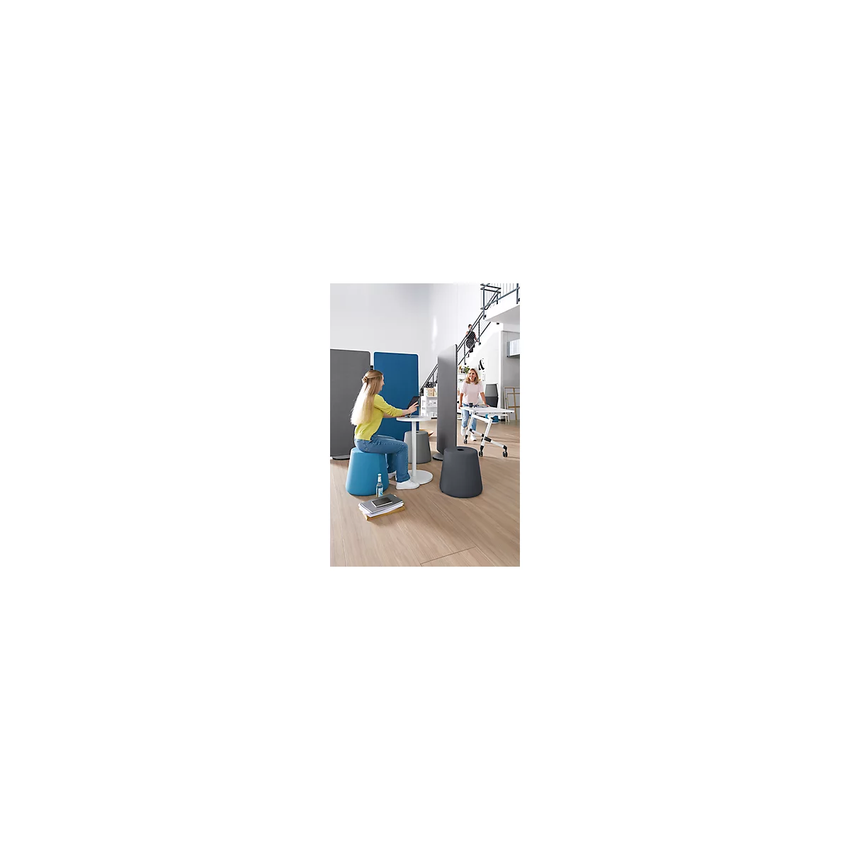 Jeune femme assise sur un tabouret bleu, travaillant sur une tablette, d'autres personnes en arrière-plan. Scène de bureau avec divers meubles.