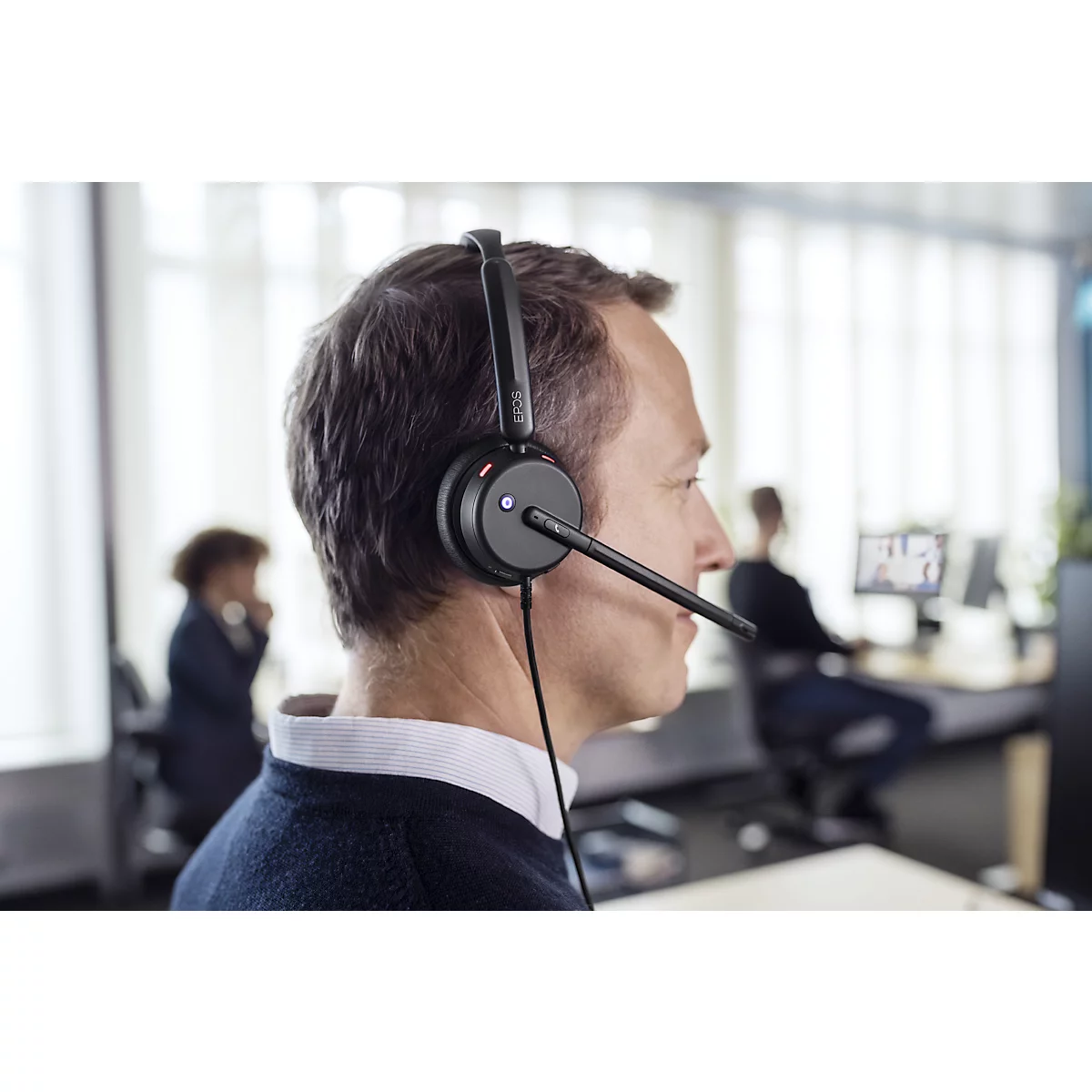 Man wearing a headset in the office. The man is wearing a dark sweater. Other people are sitting in the background.