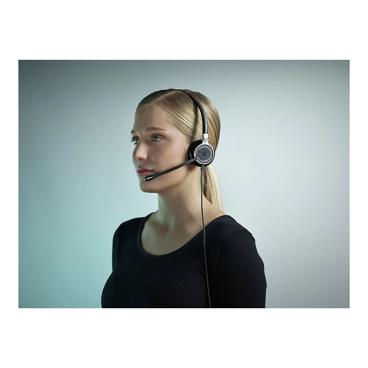 Junge Frau mit blonden Haaren trÃ¤gt ein Headset mit Mikrofon. Sie blickt zur Seite, vor hellem Hintergrund.