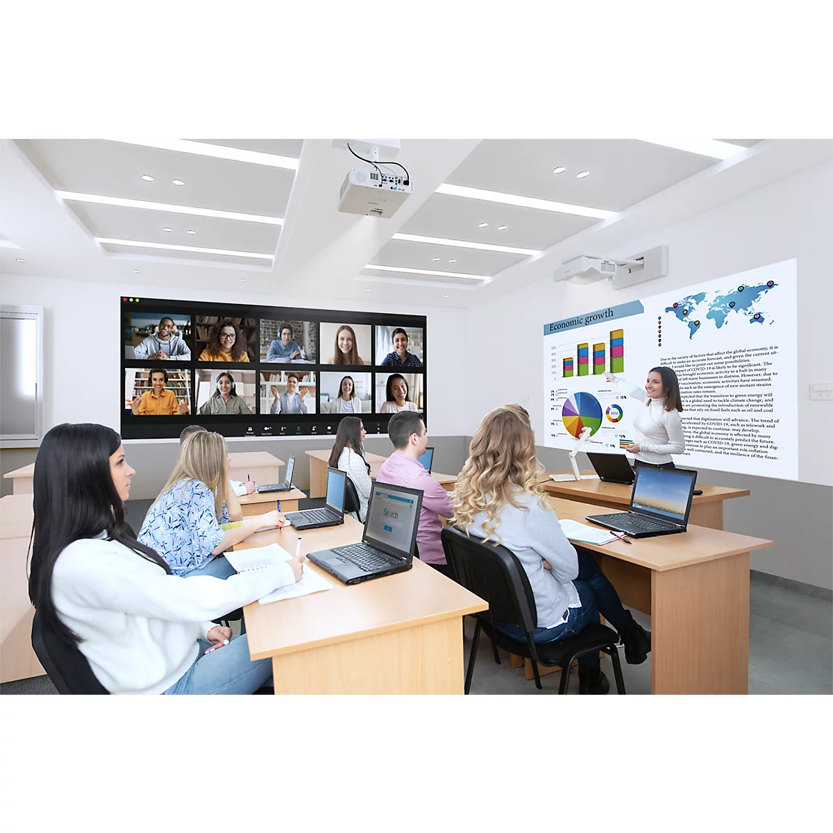 In a classroom, a woman stands in front of a screen displaying diagrams and a world map. There is a video conference on a separate screen.