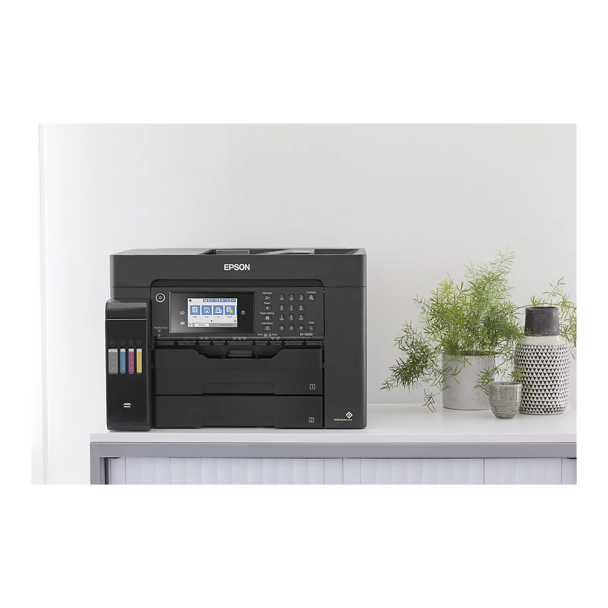 Ein schwarzer Epson-Multifunktionsdrucker steht auf einem weißen Schreibtisch. Dahinter eine weiße Wand. Auf dem Tisch sind dekorative Elemente wie Pflanzen und Vasen zu sehen.