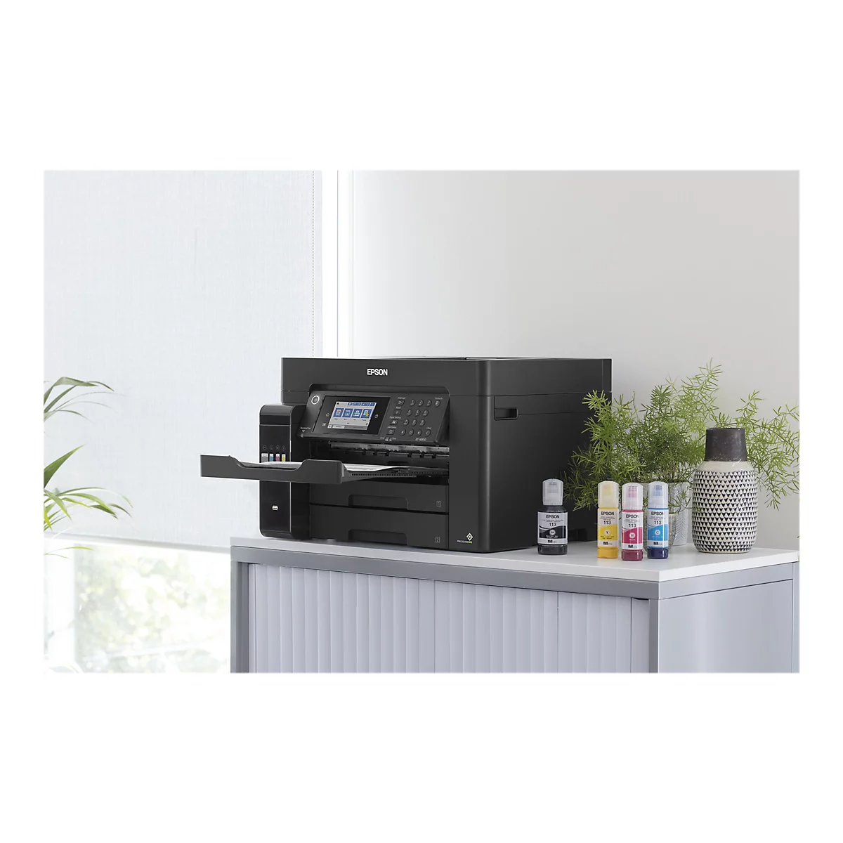 Schwarzer Epson Drucker auf einem weißen Schrank mit Tintenpatronen und Pflanzen; Fenster im Hintergrund.
