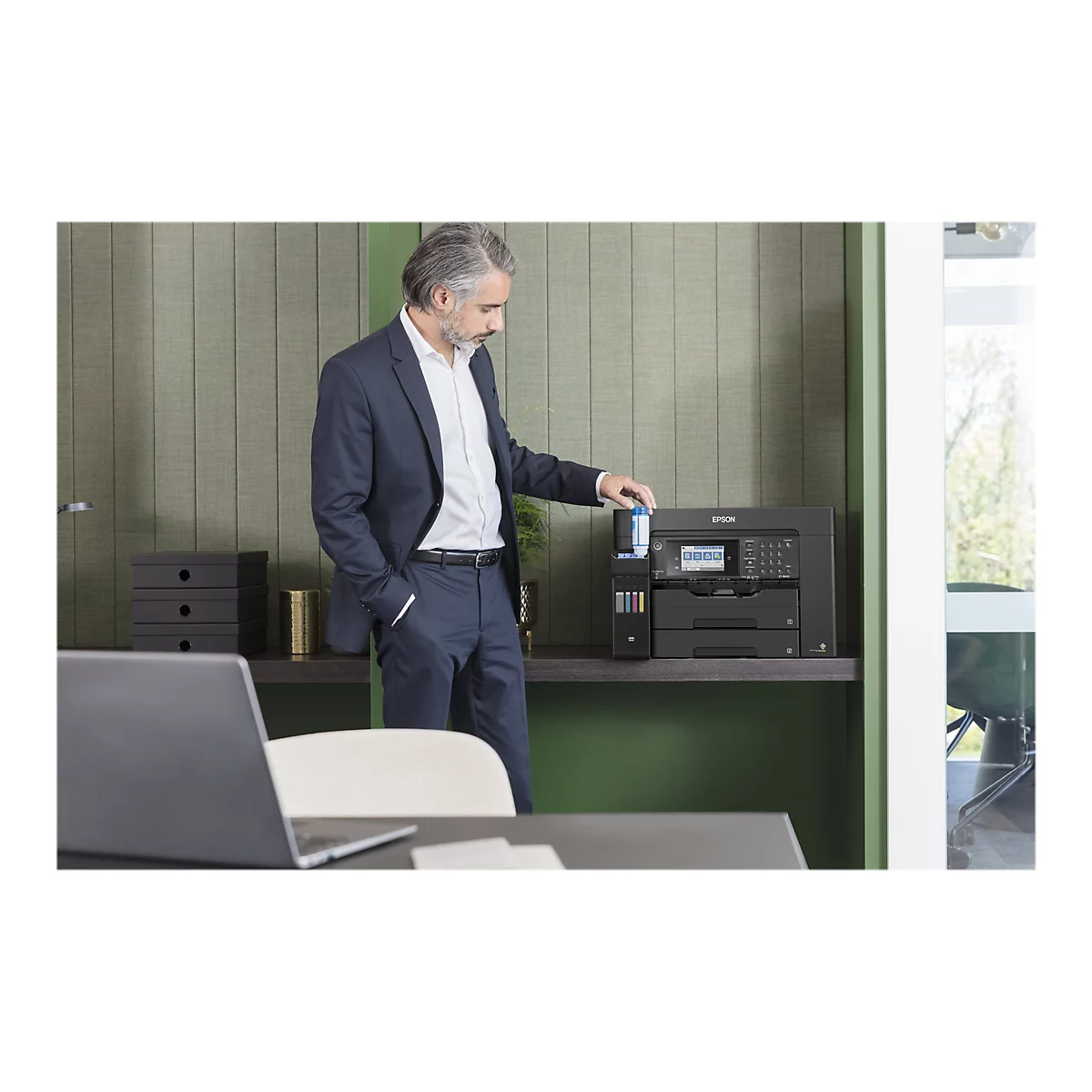 Ein Mann in Anzug befüllt einen Tintenstrahldrucker. Der Drucker steht auf einem Regal vor einer grünen Wand. Im Hintergrund ein Fenster.