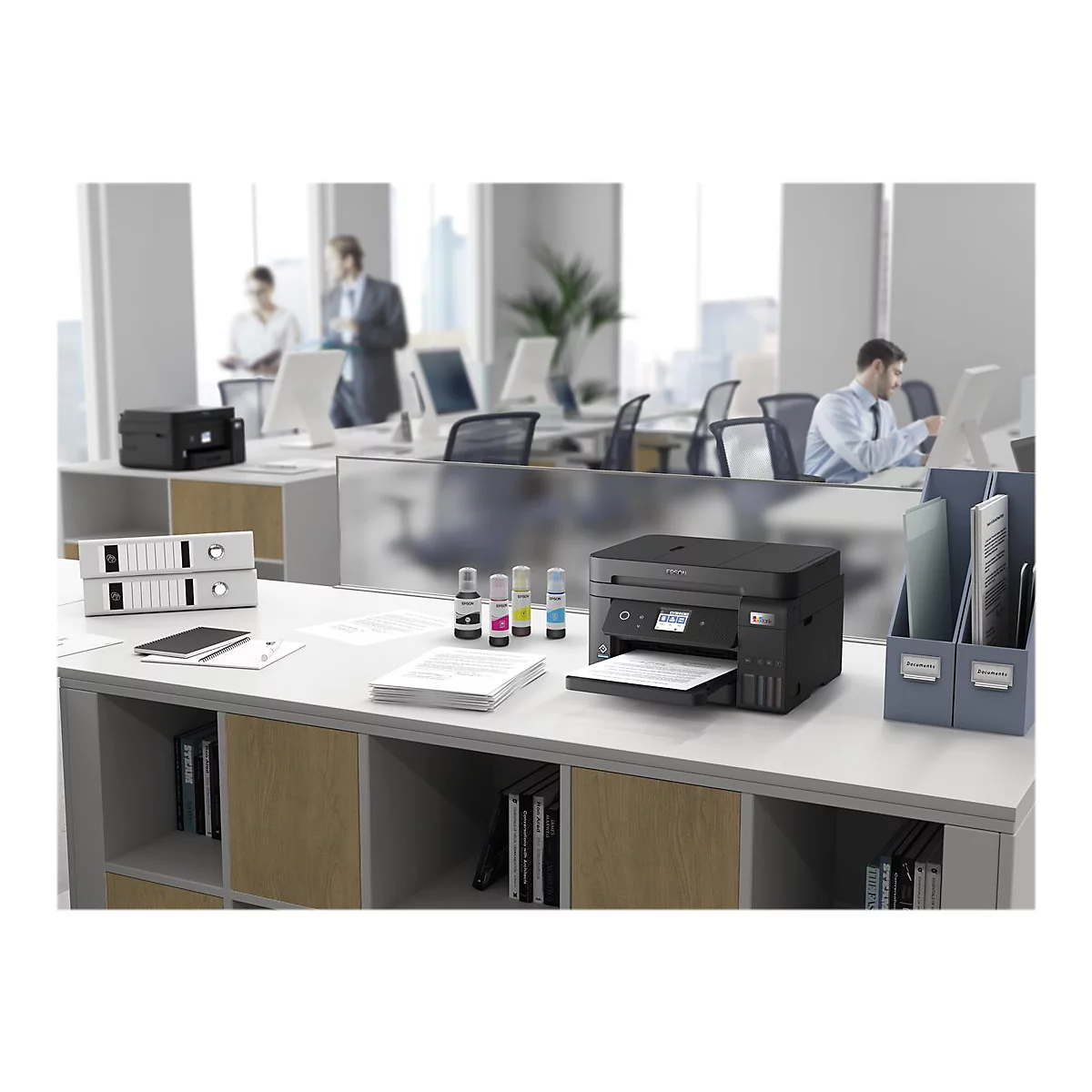 Ein Drucker steht auf einem Schreibtisch in einem BÃ¼ro. Im Hintergrund arbeiten weitere Personen.