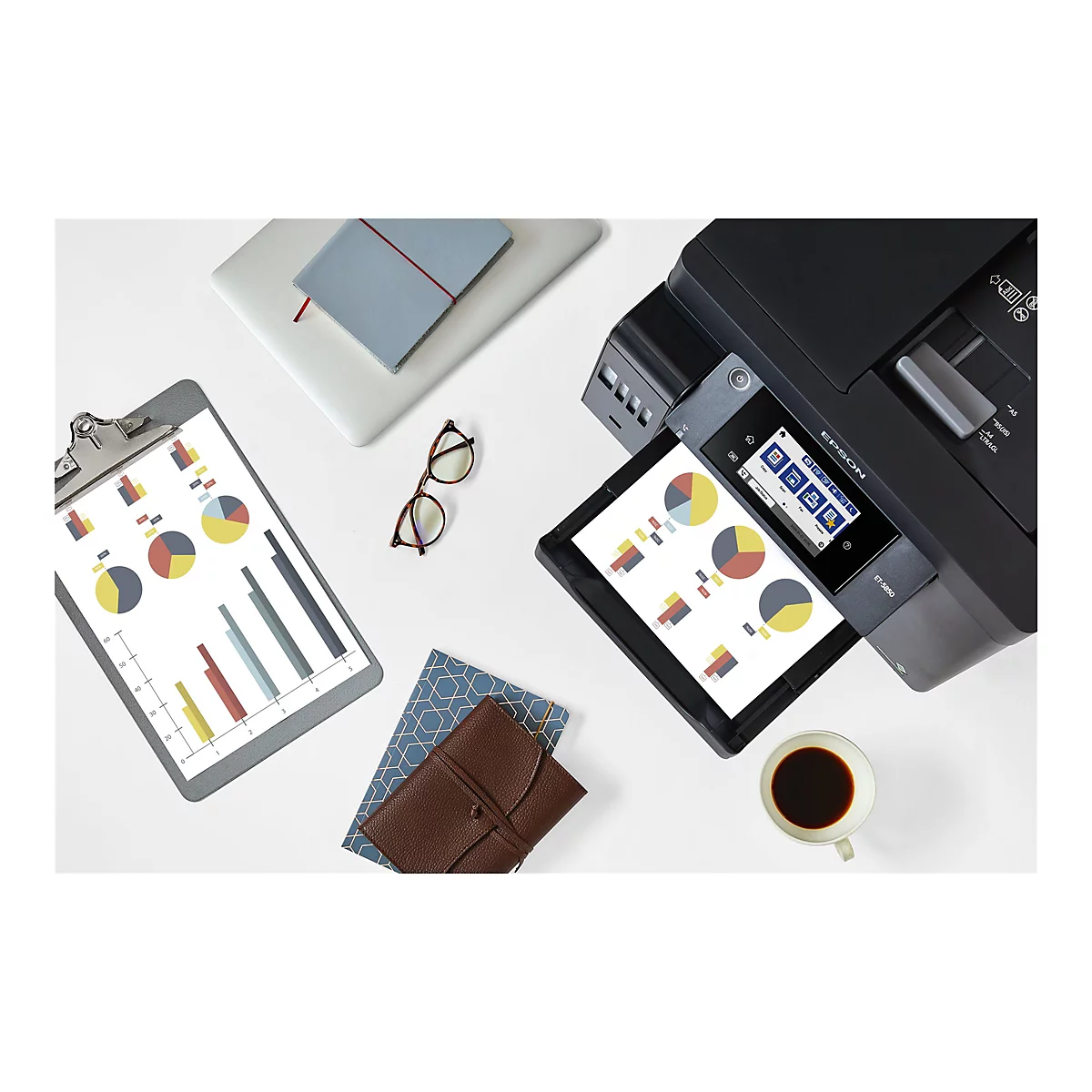 Draufsicht auf einen Schreibtisch mit Drucker, Diagrammen auf Papier, Brille, Kaffee und Notizbuch.