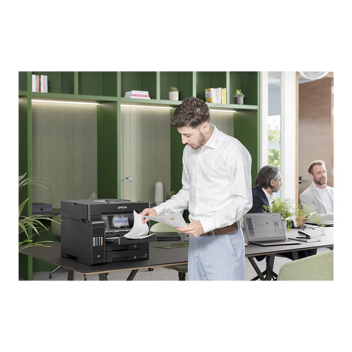 Ein Mann entnimmt Papier aus einem Epson-Drucker. Im Hintergrund sitzen zwei weitere Männer an einem Tisch.