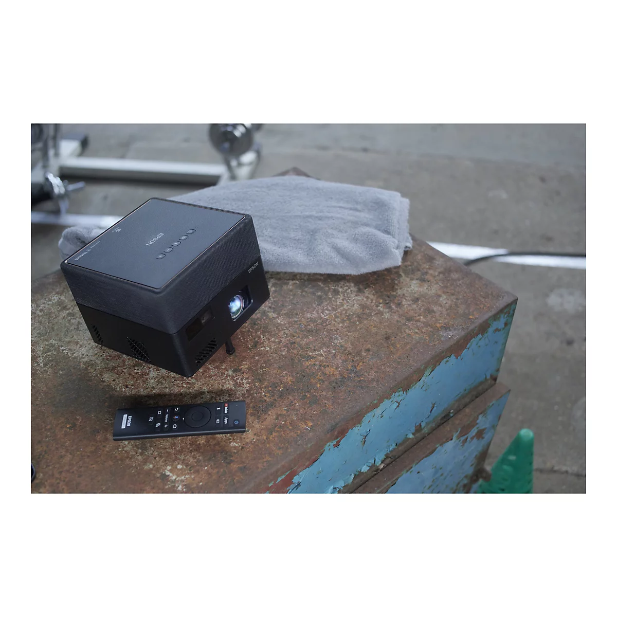 Ein schwarzer Epson-Projektor steht auf einer rostigen, blau-braun gestrichenen Metalloberfläche. Rechts liegt eine Fernbedienung, links ein graues Handtuch.