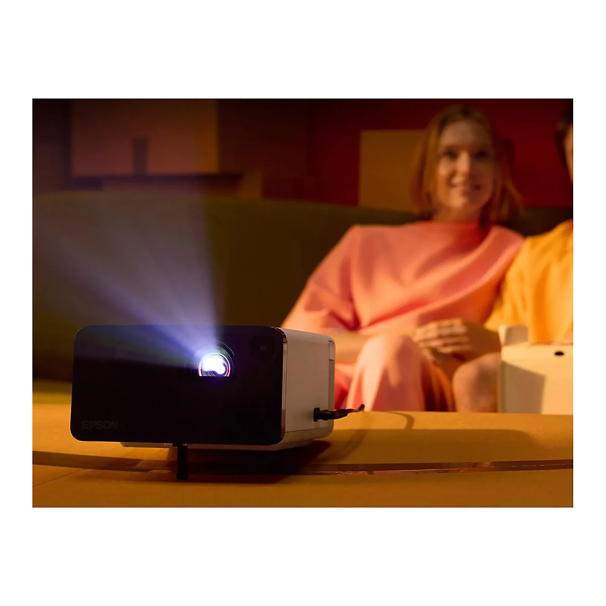 Beamer projiziert Lichtstrahl. Im Hintergrund sitzen zwei Personen auf einem Sofa.