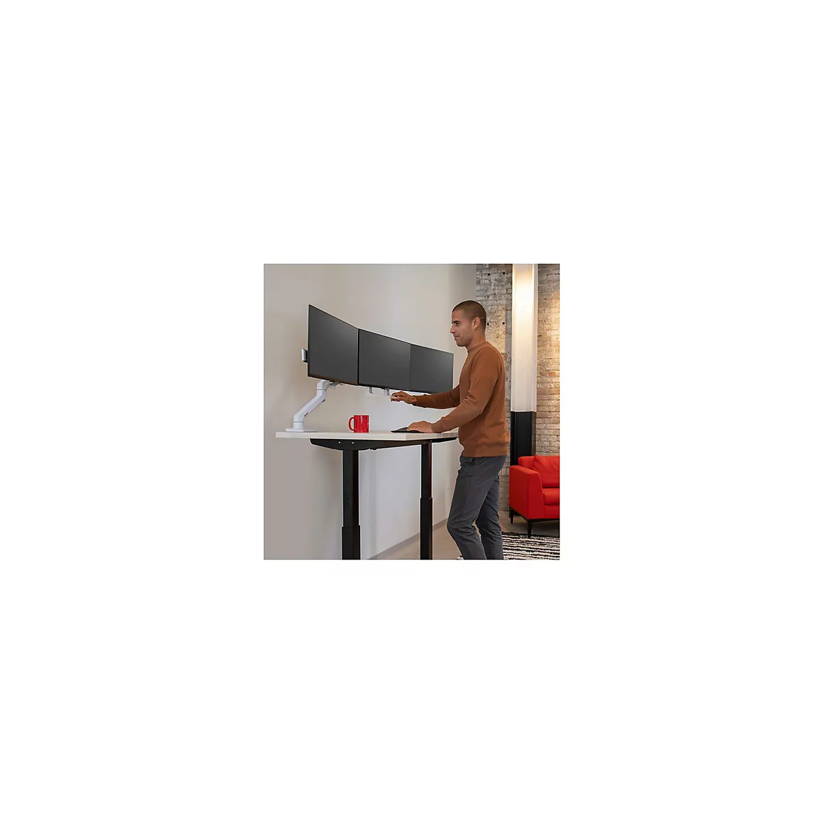 Ein Mann steht an einem höhenverstellbaren Schreibtisch und bedient drei Monitore. Auf dem Tisch steht eine rote Tasse.