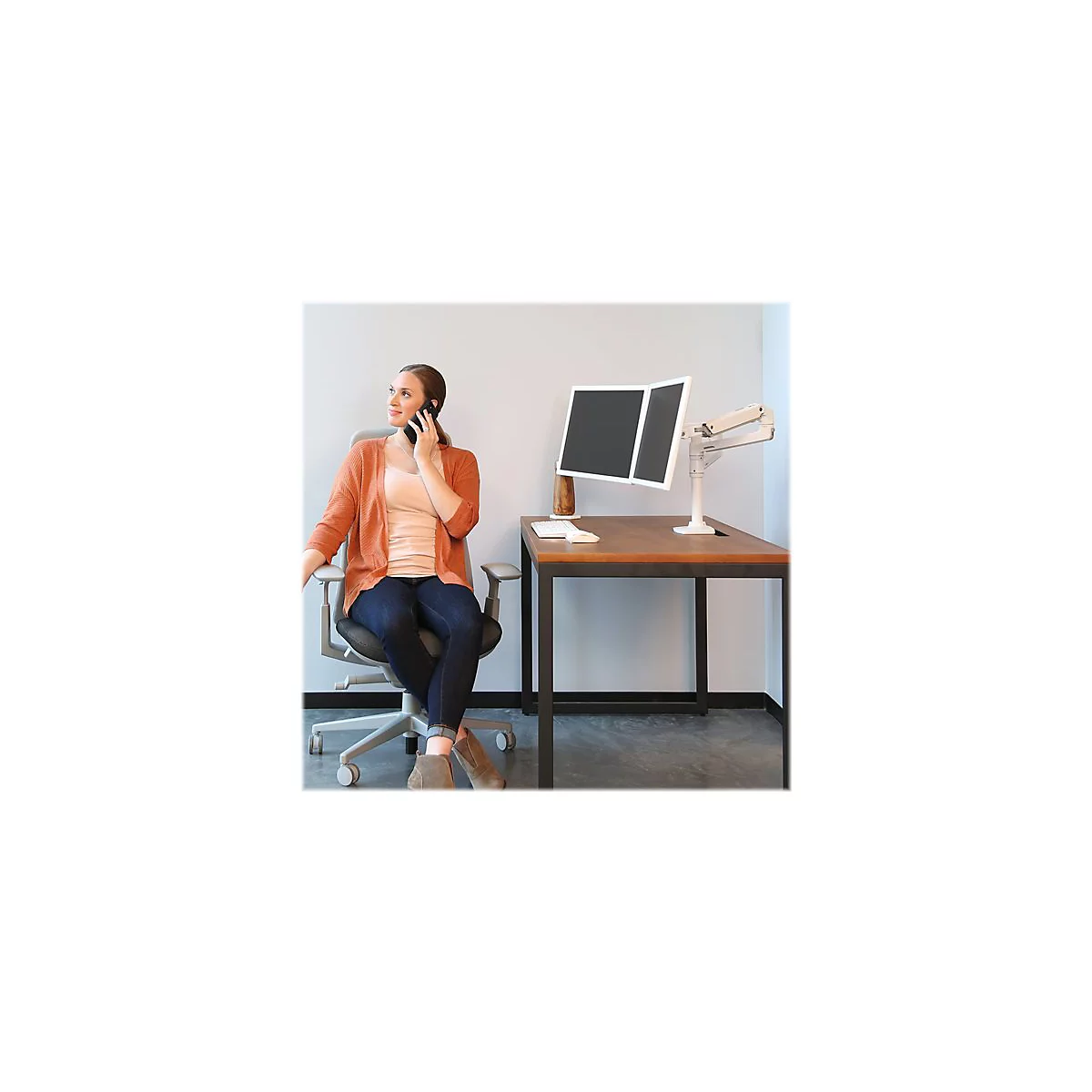 Eine Frau am Telefon sitzt an einem Schreibtisch mit zwei Bildschirmen. Sie trÃ¤gt einen orangefarbenen Cardigan, Jeans und Stiefel.