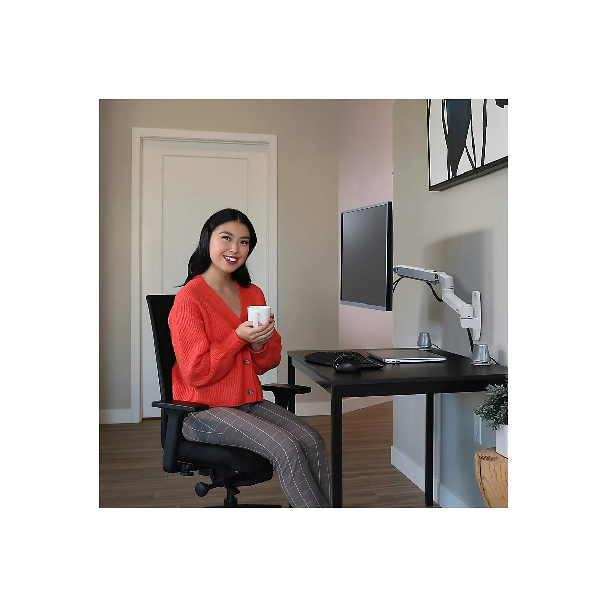 Eine Frau im roten Cardigan sitzt an einem Schreibtisch und lächelt in die Kamera, während sie eine weiße Tasse hält. Auf dem Schreibtisch ein Monitor und Laptop.