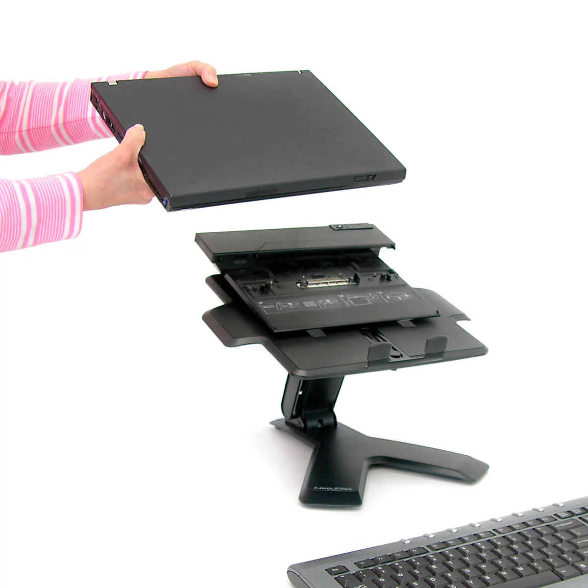 Eine Person hält ein schwarzes Laptop über eine schwarze Laptop-Dockingstation, die an einen Ständer befestigt ist, mit einer Tastatur im Hintergrund.