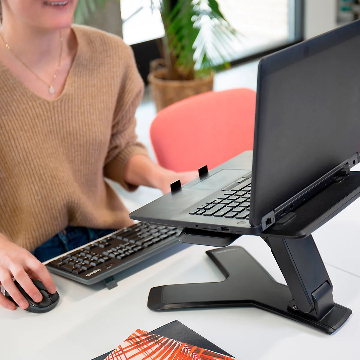 Person arbeitet an einem Laptop, der auf einem Ständer steht. Auf dem Tisch befinden sich Tastatur, Maus und Notizen.